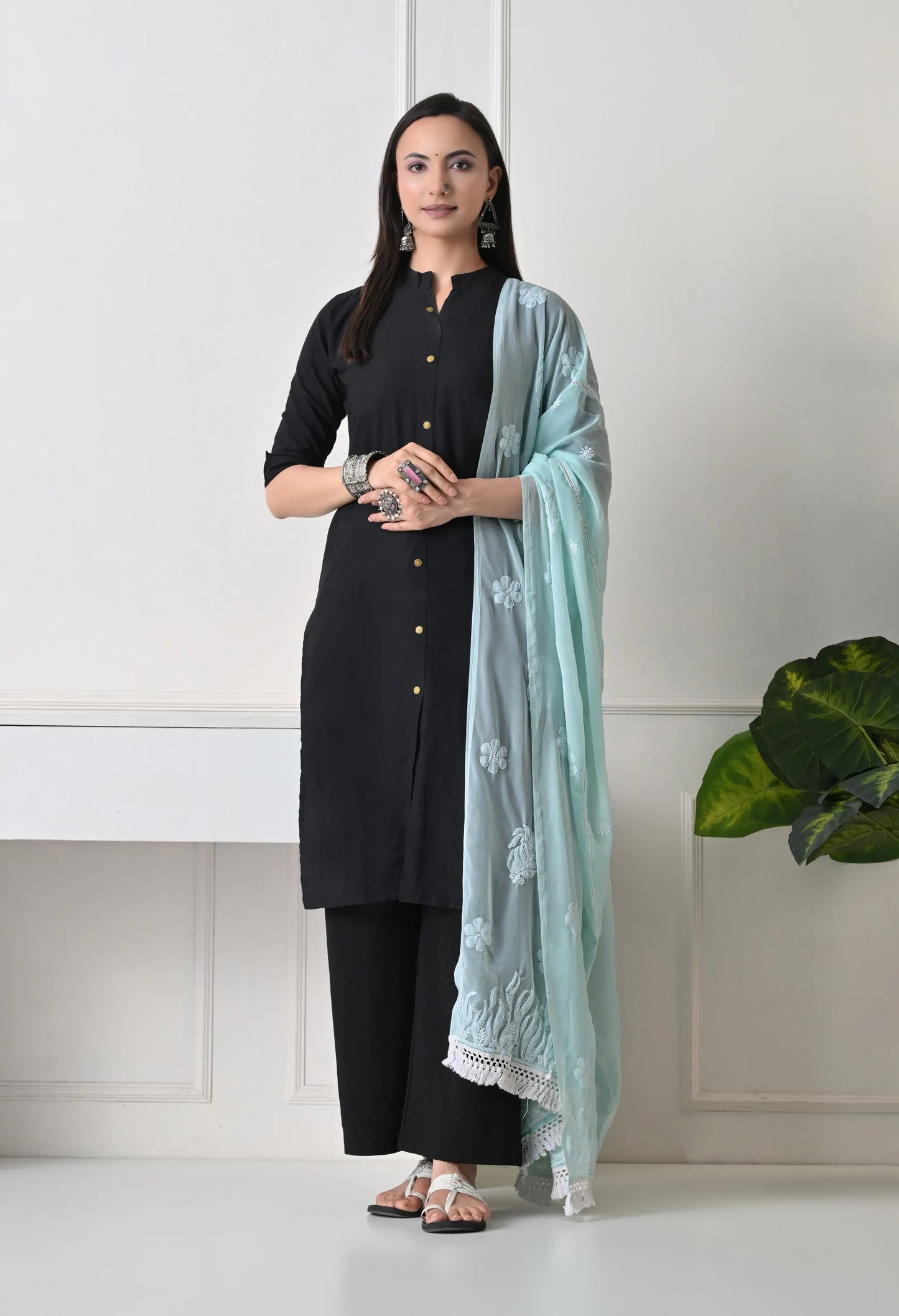 Woman in a black outfit with a mint green dupatta standing against a white wall.
